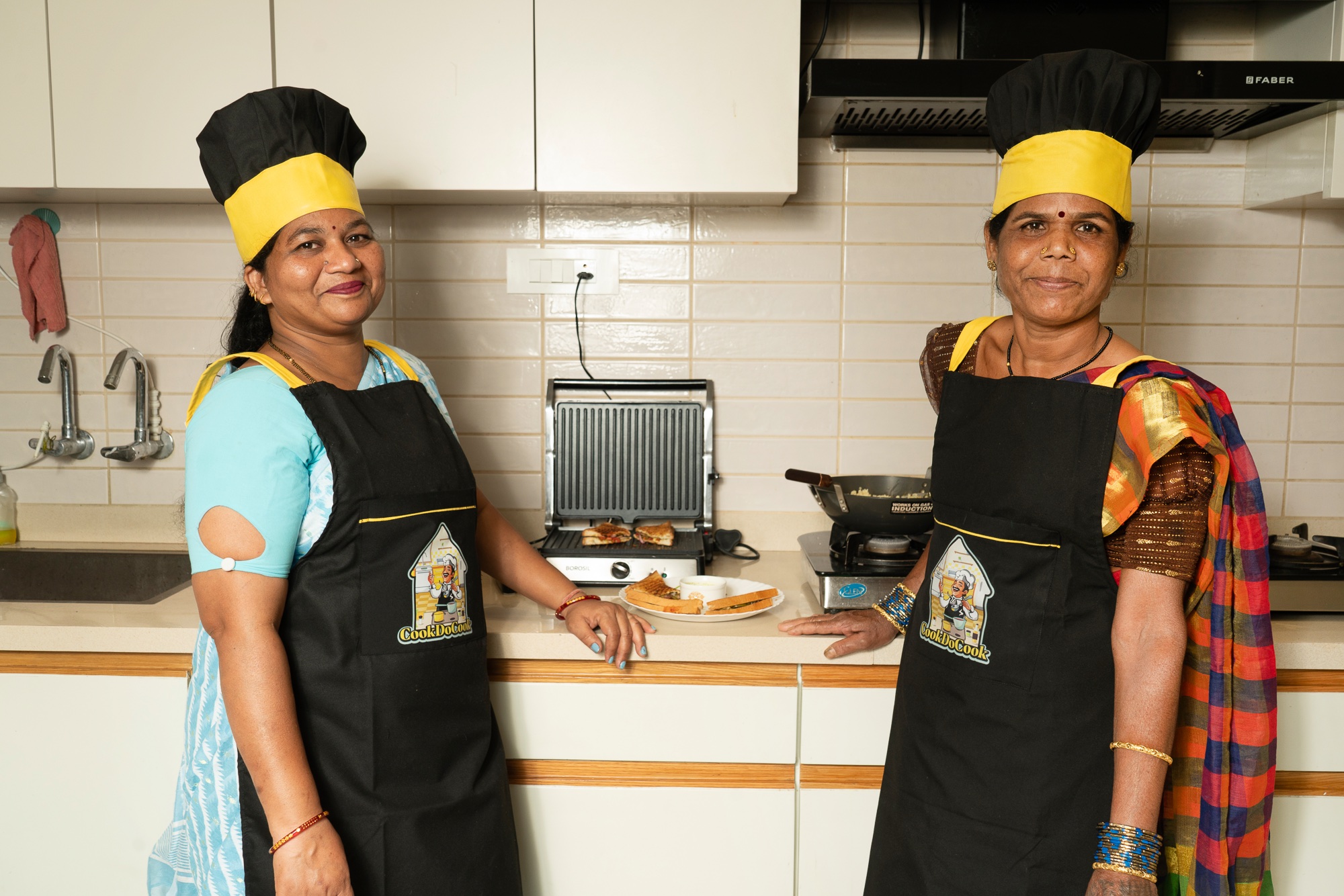 Two CookDoCook cooks standing together in a kitchen beside a sandwich press, one resting a hand on the counter.